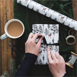 May include: A person wrapping a gift with white wrapping paper featuring a black and white forest and deer design. The wrapping paper is being tied with a black ribbon.
