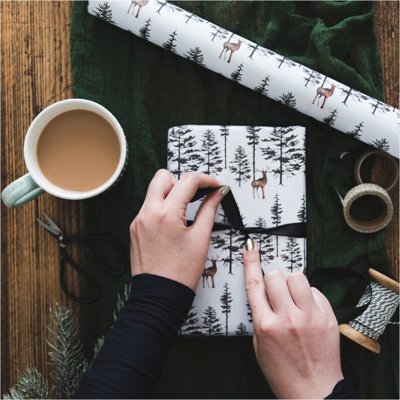 May include: A person wrapping a gift with white wrapping paper featuring a black and white forest and deer design. The wrapping paper is being tied with a black ribbon.