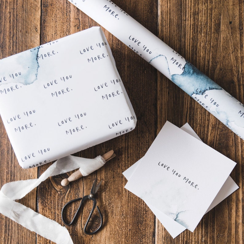 May include: White wrapping paper with the words "Love you more." repeated in a blue watercolour pattern. A roll of the wrapping paper is shown with a smaller piece of the paper on a wooden surface. A pair of scissors and a spool of white ribbon are also shown.