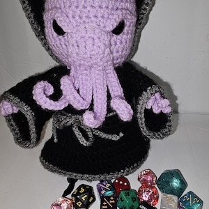 May include: A crocheted purple Cthulhu doll wearing a black robe with gray trim. The doll has black eyes and tentacles. A collection of colorful polyhedral dice are scattered in front of the doll, including a red die with the word "LIFELINK" on it.