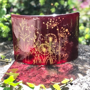 May include: A curved, claret-coloured glass vase with gold floral designs. The vase has a glossy finish and is decorated with delicate illustrations of wildflowers and grasses. The background is blurred, with green foliage.