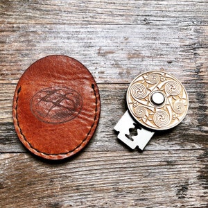 Könnte beinhalten: Ein braunes Lederetui mit einem genähten Rand und einem Design eines geflochtenen Knotens. Daneben liegt ein Holzrasierer mit einer silbernen Klinge und einem Design eines keltischen Knotens.