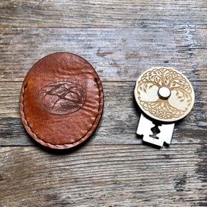 Könnte beinhalten: Ein braunes Lederetui mit einem genähten Rand und einem Design einer Bergkette. Daneben liegt ein Holzrasierer mit einem Lebensbaum-Design und einer silbernen Klinge.