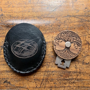 Puede incluir: Una bolsa de cuero negro con un cierre de presión de metal plateado y un diseño detallado de dos pájaros en vuelo. Junto a la bolsa hay una navaja circular de madera con un diseño de árbol de la vida y una cuchilla de metal plateado.