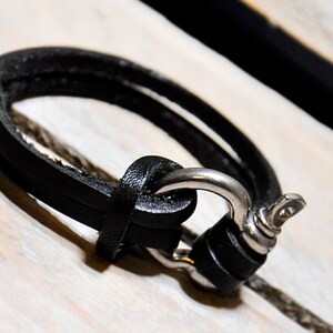Puede incluir: Un brazalete de cuero negro con un cierre de plata. El brazalete es de doble capa y tiene un diseño simple.
