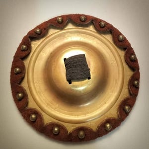 May include: A round, gold-colored metal jingle with a brown leather trim and a black elastic band. The jingle has a scalloped edge and is decorated with small, round metal studs.