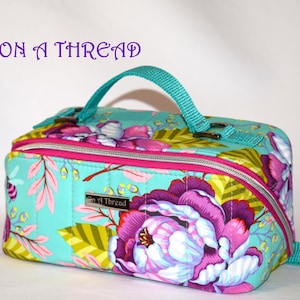 Puede incluir: Bolso rectangular con estampado floral, con fondo turquesa y grandes flores moradas y rosas. Tiene un asa y cremallera turquesa, con el texto "ON a THREAD" encima. La cremallera est&aacute; delineada en rosa.