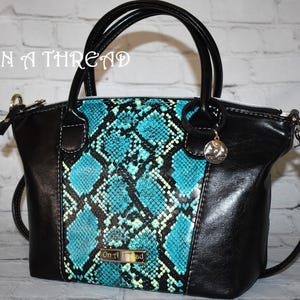 May include: A black and turquoise snakeskin-patterned handbag with a top handle and a detachable shoulder strap. The bag has a gold-colored zipper and a small metal charm with the words "On a Thread" on it.