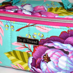 Puede incluir: Bolso con estampado floral con fondo turquesa, con grandes flores moradas y rosas, detalles en rosa y cremallera plateada. El bolso tiene una correa turquesa y el texto "On A Thread" en una placa plateada.