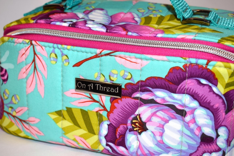 Puede incluir: Bolso con estampado floral con fondo turquesa, con grandes flores moradas y rosas, detalles en rosa y cremallera plateada. El bolso tiene una correa turquesa y el texto "On A Thread" en una placa plateada.