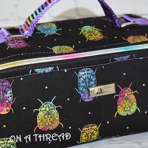 May include: Black zippered travel case with a rainbow beetle print and the text "On A Thread" on the bottom. The case has a purple zipper pull and a silver zipper.