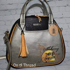 May include: A silver and black handbag with a yellow tassel and a skull design embroidered on the front. The bag has a black strap and a yellow zipper. The text "Alchemy" is embroidered above the skull.