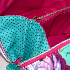 Puede incluir: Vista abierta de un bolso colorido con un estampado floral y de abejas. El bolso presenta un exterior turquesa con detalles florales rosas y morados, un interior turquesa con lunares azules y un forro rosa brillante. Se ve una cremallera plateada.