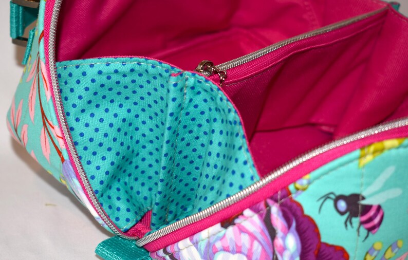 Puede incluir: Vista abierta de un bolso colorido con un estampado floral y de abejas. El bolso presenta un exterior turquesa con detalles florales rosas y morados, un interior turquesa con lunares azules y un forro rosa brillante. Se ve una cremallera plateada.