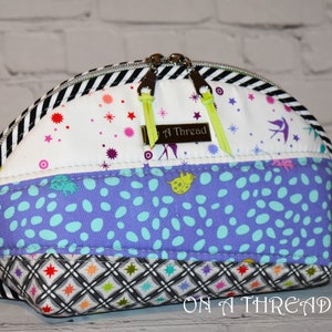 May include: A small, half-circle shaped zippered pouch with a black and white striped border. The pouch is made of colorful patterned fabric with a label that reads "On A Thread".