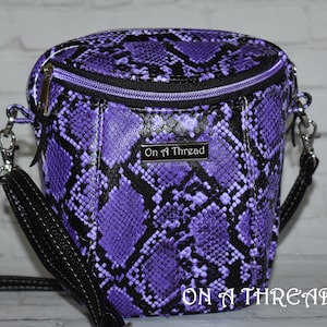 May include: Purple and black snakeskin patterned crossbody bag with a black strap and a zipper closure. The bag has a silver metal tag that reads "On a Thread".