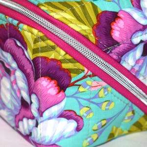 Puede incluir: Primer plano de un neceser con estampado floral. La bolsa presenta un dise&ntilde;o vibrante con grandes flores moradas y rosas sobre un fondo turquesa. Una cremallera rosa brillante con un tirador plateado a&ntilde;ade un toque de color.