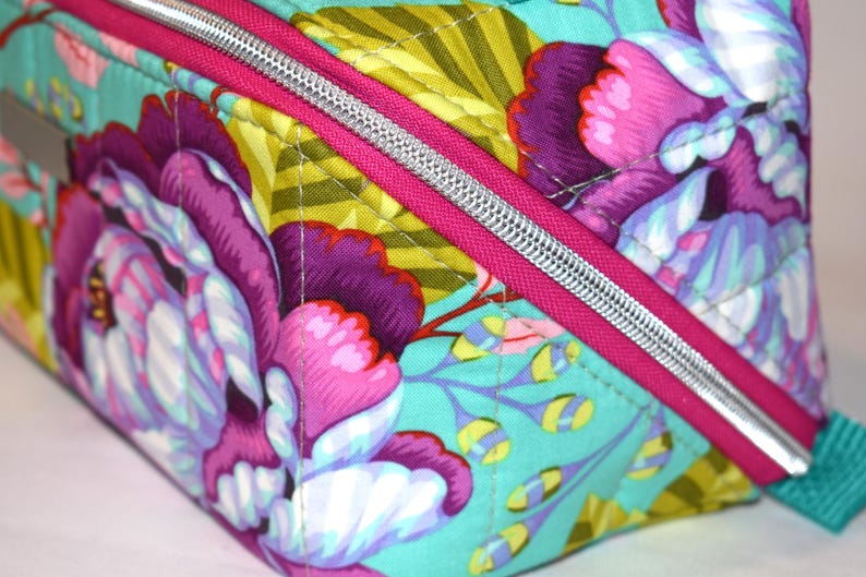 Puede incluir: Primer plano de un neceser con estampado floral. La bolsa presenta un dise&ntilde;o vibrante con grandes flores moradas y rosas sobre un fondo turquesa. Una cremallera rosa brillante con un tirador plateado a&ntilde;ade un toque de color.