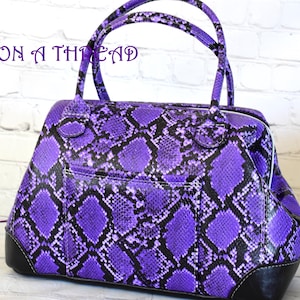 May include: A purple handbag with a snakeskin pattern and black accents. The bag has two handles and a pocket on the front. The text "ON A THREAD" is written on the top left of the bag.