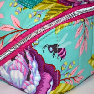 Puede incluir: Primer plano de un art&iacute;culo de tela acolchada con un dise&ntilde;o floral y de abeja. La tela es de color verde azulado claro con estampados florales rosas, morados y amarillos. Se ve una cremallera plateada con un borde rosa brillante. Hay una peque&ntilde;a abeja.