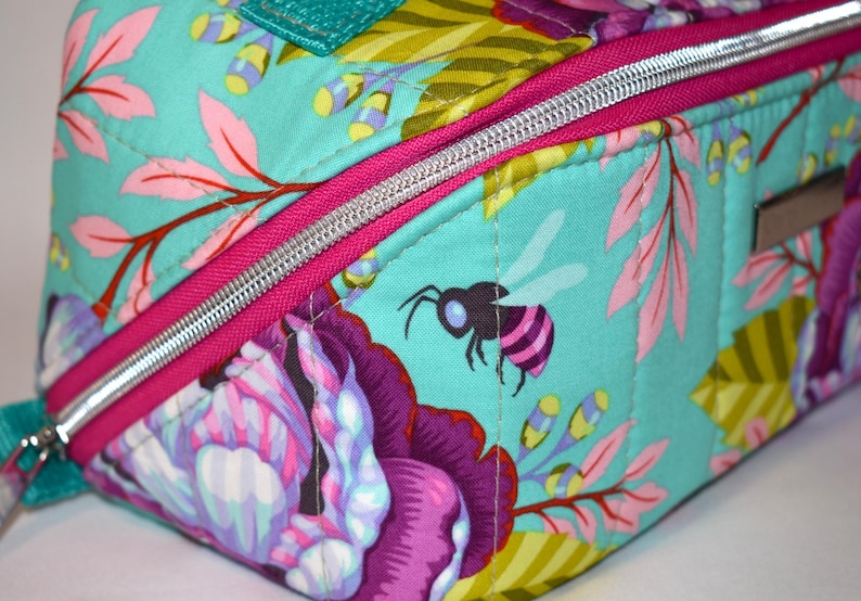 Puede incluir: Primer plano de un art&iacute;culo de tela acolchada con un dise&ntilde;o floral y de abeja. La tela es de color verde azulado claro con estampados florales rosas, morados y amarillos. Se ve una cremallera plateada con un borde rosa brillante. Hay una peque&ntilde;a abeja.