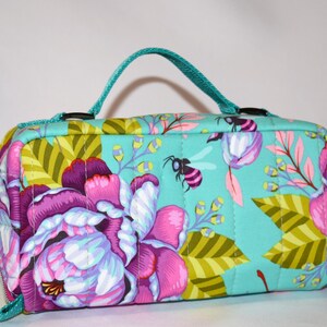 Puede incluir: Un neceser rectangular acolchado con fondo turquesa y estampado floral en tonos rosas, morados y verdes. El neceser tiene un asa turquesa, cierre de cremallera y dise&ntilde;o de abeja.