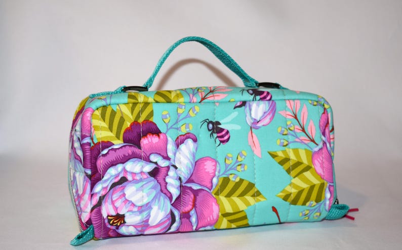 Puede incluir: Un neceser rectangular acolchado con fondo turquesa y estampado floral en tonos rosas, morados y verdes. El neceser tiene un asa turquesa, cierre de cremallera y dise&ntilde;o de abeja.