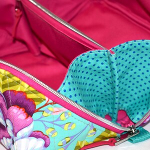 Puede incluir: Vista abierta de un neceser con estampado floral y un interior rosa brillante. La bolsa presenta un exterior turquesa con grandes flores moradas y rosas, detalles amarillos y un bolsillo interior de lunares turquesa. Una cremallera plateada recorre el borde.