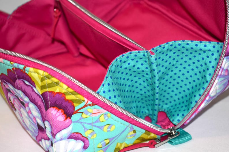 Puede incluir: Vista abierta de un neceser con estampado floral y un interior rosa brillante. La bolsa presenta un exterior turquesa con grandes flores moradas y rosas, detalles amarillos y un bolsillo interior de lunares turquesa. Una cremallera plateada recorre el borde.
