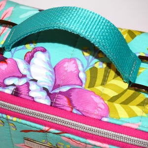 Puede incluir: Bolso de tela acolchada con fondo turquesa y estampado floral en rosa, morado y amarillo. Cuenta con un asa y correas turquesas con hebillas met&aacute;licas marrones. Una cremallera y ribete rosas acent&uacute;an el dise&ntilde;o.