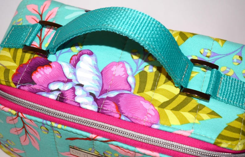Puede incluir: Bolso de tela acolchada con fondo turquesa y estampado floral en rosa, morado y amarillo. Cuenta con un asa y correas turquesas con hebillas met&aacute;licas marrones. Una cremallera y ribete rosas acent&uacute;an el dise&ntilde;o.