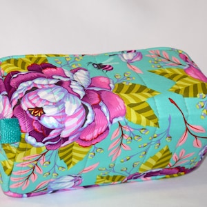 Puede incluir: Un neceser rectangular con estampado floral. La bolsa presenta grandes flores rosas y moradas, hojas verdes y un fondo turquesa. Tiene asas turquesas y cierre de cremallera. La bolsa mide aproximadamente 20 cm de largo y 13 cm de alto.