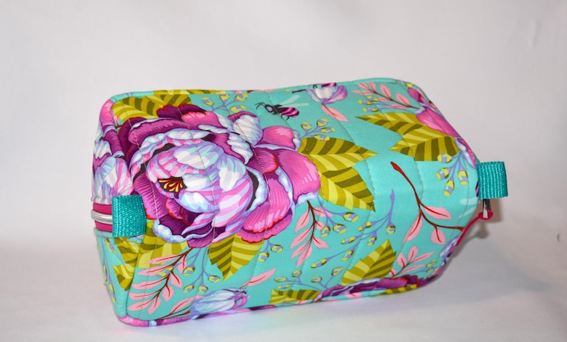 Puede incluir: Un neceser rectangular con estampado floral. La bolsa presenta grandes flores rosas y moradas, hojas verdes y un fondo turquesa. Tiene asas turquesas y cierre de cremallera. La bolsa mide aproximadamente 20 cm de largo y 13 cm de alto.
