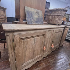May include: A light-colored wooden sideboard with two doors and decorative hardware. The doors have a carved design and are adorned with peach-colored tassels. A painting and a wicker basket sit on top of the sideboard.