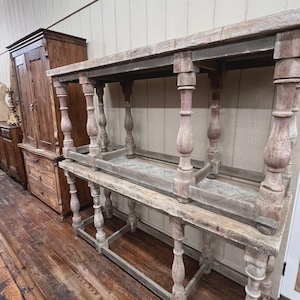 May include: Two-tiered, antique wooden console table with turned legs and a distressed finish. The table has a rectangular top and a lower shelf, showcasing a rustic, vintage aesthetic. The wood has a weathered, gray-brown color.