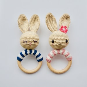 Op de afbeelding: Twee gehaakte konijnen rammelaars met houten ringen. Eén konijn draagt een blauw-wit gestreept shirt en de andere draagt een roze-wit gestreept shirt.