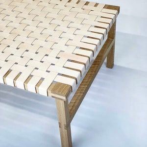 May include: A white leather woven bench with a natural wood frame. The bench has a simple design with four legs and a rectangular top.