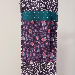 May include: A quilted fabric item with a floral pattern in shades of purple, pink, and teal. The item features a pocket with a teal polka dot fabric accent. The edges are trimmed with a white floral design on a purple background.