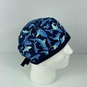 May include: A navy blue surgical scrub cap featuring a geometric shark pattern in shades of blue and white. The cap has a dark blue trim and ties at the back. The cap is designed to be worn in a medical setting.