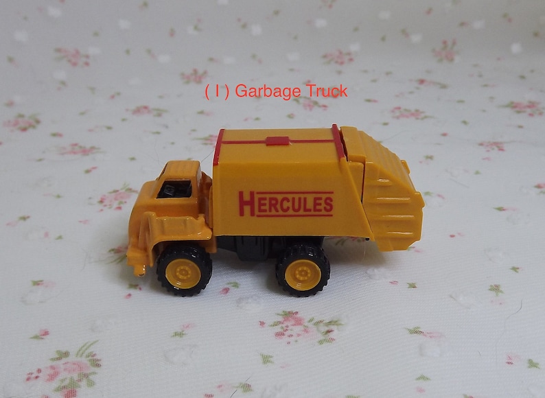 May include: Yellow toy garbage truck with a red stripe and the word "HERCULES" printed on the side.