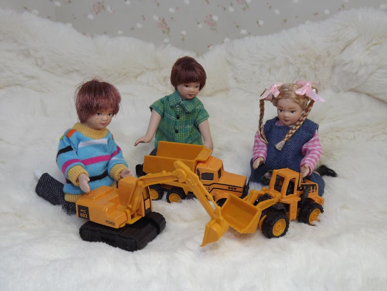 May include: Three children play with toy construction vehicles. A girl in a blue and white dress holds a yellow toy dump truck. A boy in a blue and pink striped shirt holds a yellow toy excavator. A girl in a green and white plaid shirt holds a yellow toy dump truck.