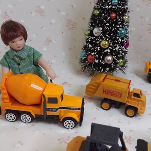 May include: A collection of yellow toy construction vehicles, including a cement mixer, a dump truck labeled "Hercules", an excavator, and a bulldozer. A doll wearing a green plaid shirt is standing next to the cement mixer.