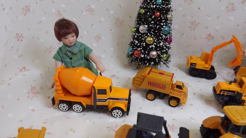 May include: A collection of yellow toy construction vehicles, including a cement mixer, a dump truck labeled "Hercules", an excavator, and a bulldozer. A doll wearing a green plaid shirt is standing next to the cement mixer.
