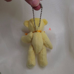 May include: A yellow plush teddy bear keychain with a silver chain and a yellow ribbon around its neck. The ribbon has the text "Love is..." printed on it.
