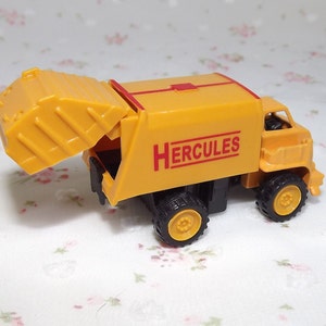 May include: Yellow toy garbage truck with a red "HERCULES" logo on the side. The truck has a lifting bin that is open.