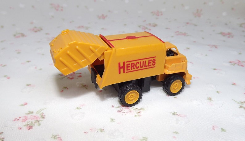 May include: Yellow toy garbage truck with a red "HERCULES" logo on the side. The truck has a lifting bin that is open.