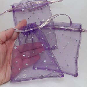 Pode incluir: Três sacos de presente de tule roxo com glitter prateado. Os sacos são feitos de um tecido fino e delicado e têm um fecho de cordão.