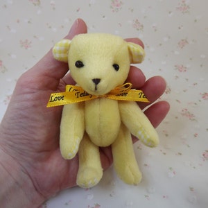 May include: A small yellow teddy bear with white gingham ears and paws. The bear is wearing a yellow ribbon with the words "Love Teddy" printed on it.
