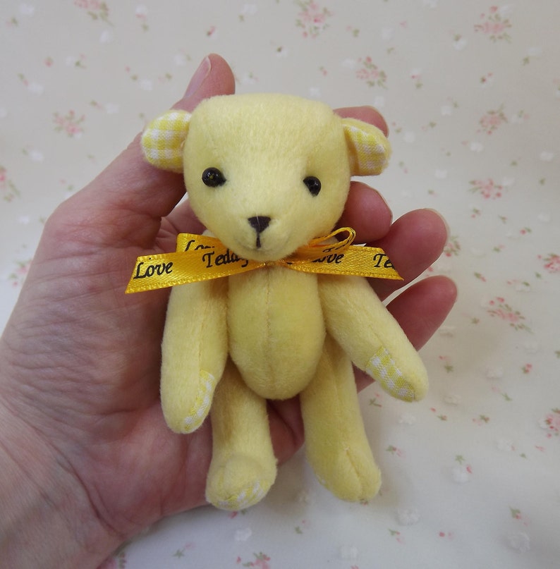 May include: A small yellow teddy bear with white gingham ears and paws. The bear is wearing a yellow ribbon with the words "Love Teddy" printed on it.