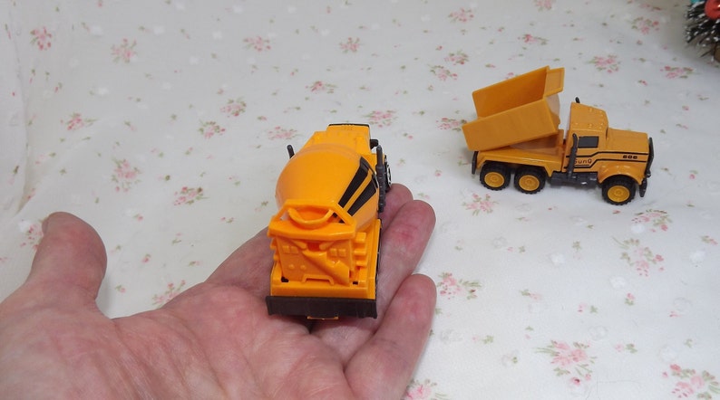 May include: Two toy construction trucks, one is a yellow dump truck with a raised bed and the other is a yellow cement mixer truck.